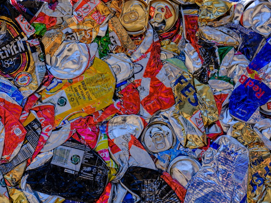 An outdoor scene showing large stacks of cardboard fruit and vegetable boxes, primarily made of brown and white corrugated material with colorful printed labels, arranged on a paved surface. Behind th