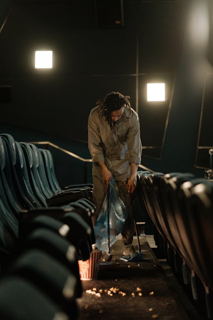 Close-up of a person wearing orange protective overalls and white gloves, holding a large blue plastic rubbish bag with their right hand. The bag appears to be filled with waste and is gathered at the