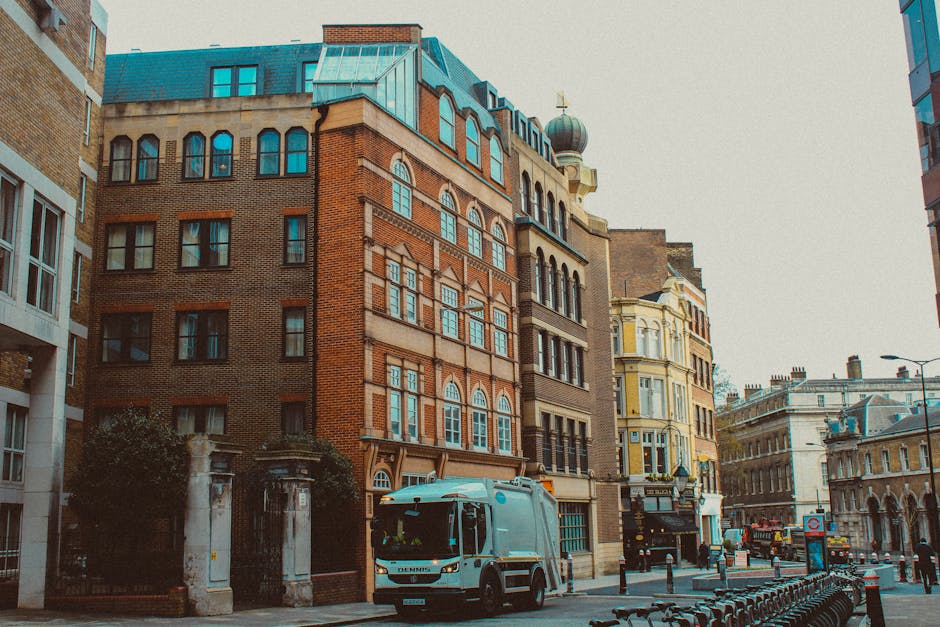 The image depicts a cityscape featuring a historic red brick and sandstone building in the foreground, with detailed masonry work, sash windows, and decorative stonework along the roofline. Behind it,