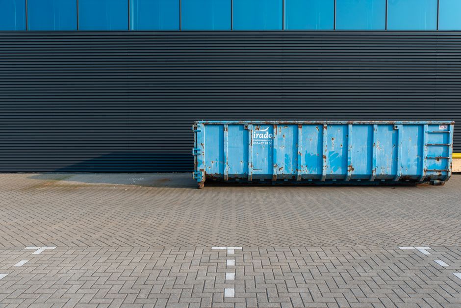 A blue metal waste container with visible rust and peeling paint, positioned on a paved surface in front of a modern building with dark horizontal cladding and a section of blue-tinted glass windows a