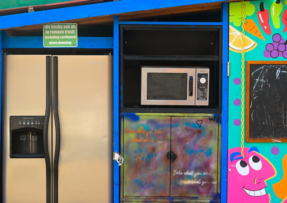 A blue metal waste container with visible rust and peeling paint, positioned on a paved surface in front of a modern building with dark horizontal cladding and a section of blue-tinted glass windows a