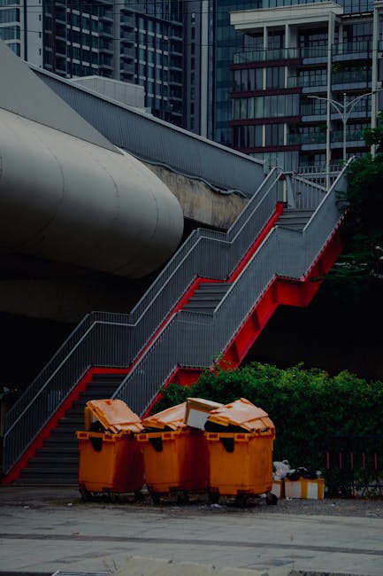 A street scene of the rear side of a multi-storey building featuring an external metal staircase with black railings leading from the ground to the upper floors, casting shadows on the peach and light